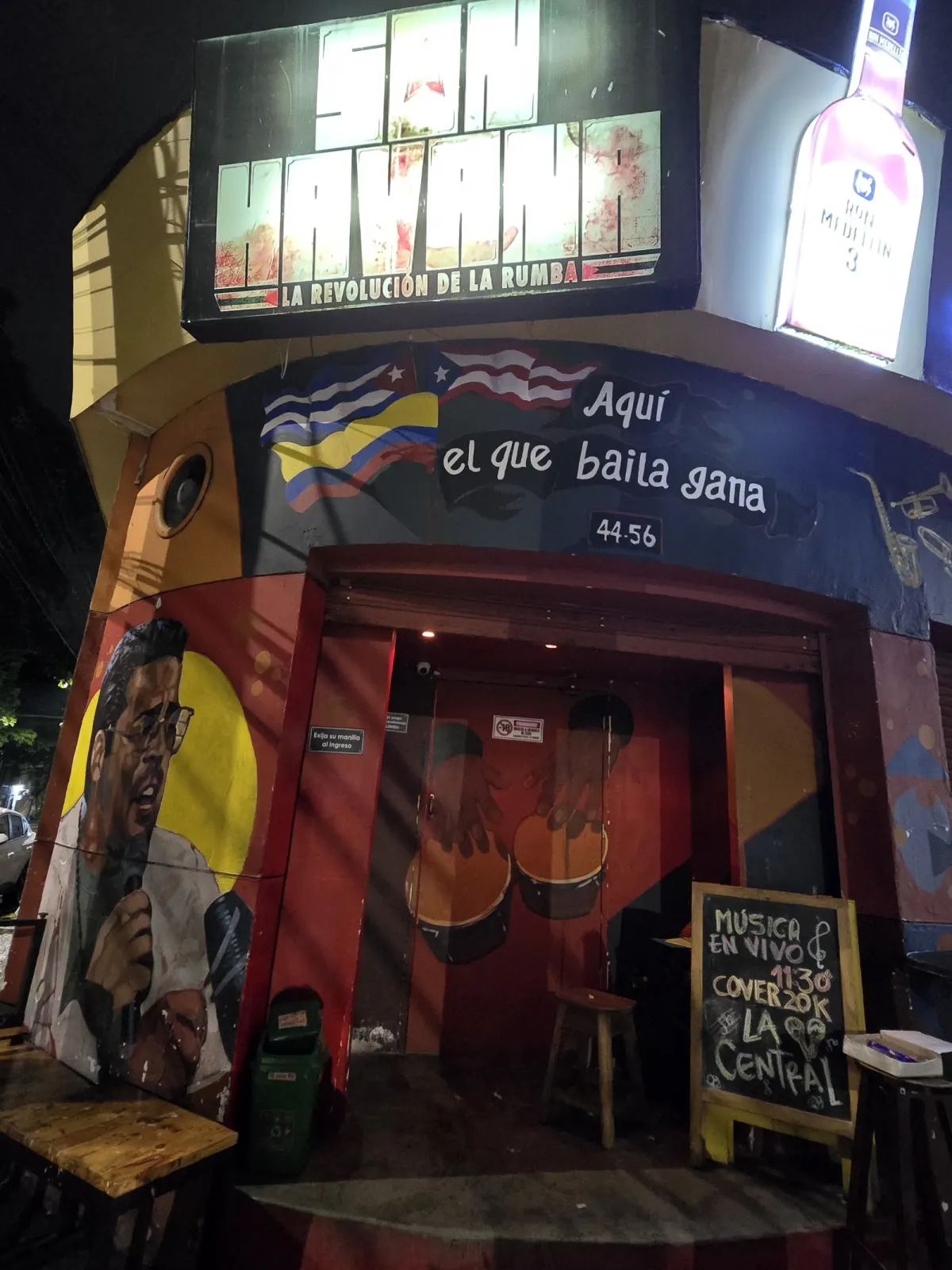 Son Havana's corner entrance at night under the sign 'La revolución de la rumba', with a painted mural of a singer and Cuban, Puerto Rican and Colombian flags beside the slogan 'Aquí el que baila gana'.