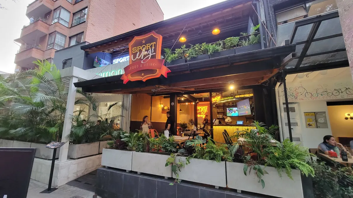 Street front of Sport Wings Laureles with its orange shield sign, a planted terrace and diners at tables inside under warm lights
