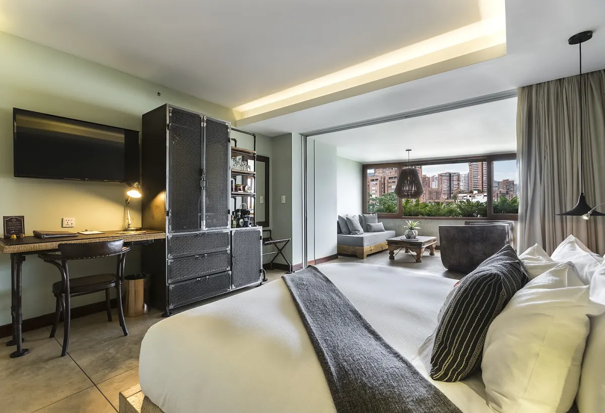 A Charlee room with a large bed, a desk, a tall metal wardrobe and a sitting area by a window looking out over El Poblado