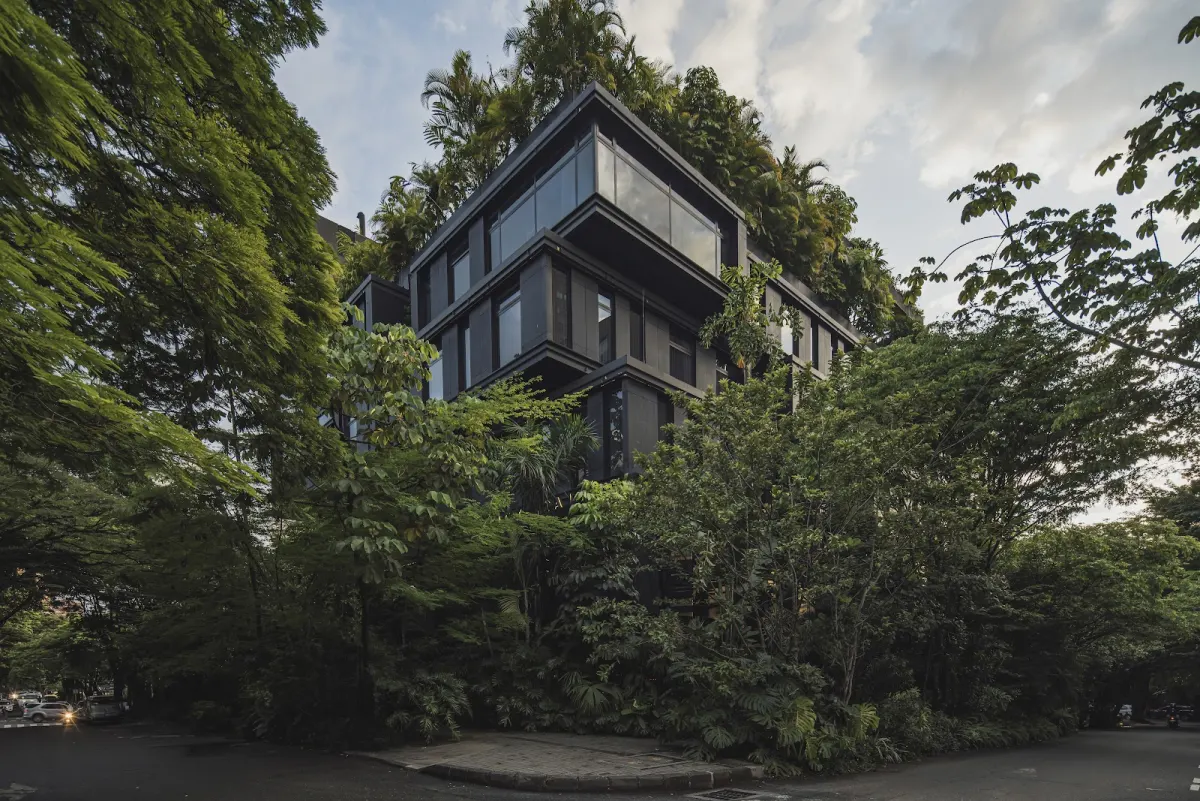 The dark, stacked facade of The Click Clack Hotel Medellín, its roof planted with palms and its corner wrapped in tall street trees