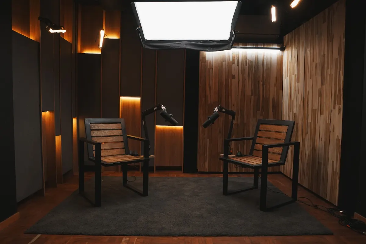 Two wooden armchairs facing each other on a dark rug at The Zone, each with a podcast microphone on a boom arm, under a large softbox and warm strip lighting on wood-panelled walls