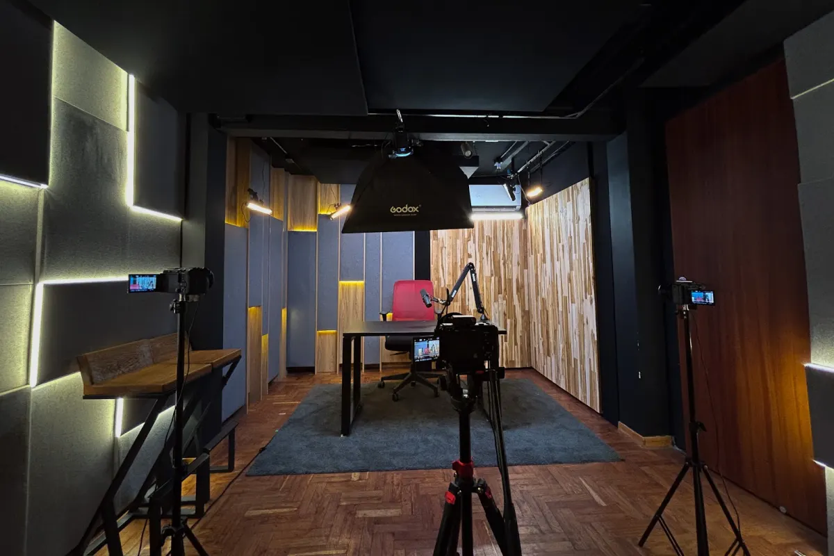 The Zone's recording room seen from behind three cameras on tripods, with a desk, a red chair and a boom-arm microphone under a Godox softbox, between felt acoustic panels and wood-slat walls