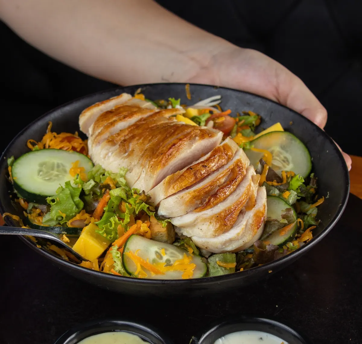 Grilled chicken breast sliced over a salad of lettuce, cucumber, mango and grated cheese in a black bowl at Vibe Medellín