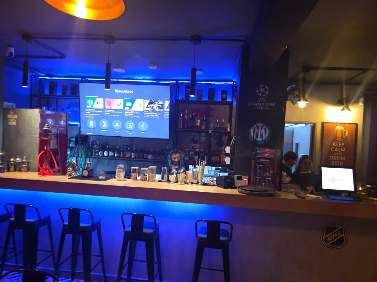 Blue-lit bar counter at Vibe Medellín with metal stools, a TV above the back bar, a hookah on the counter and a Champions League logo on the pillar