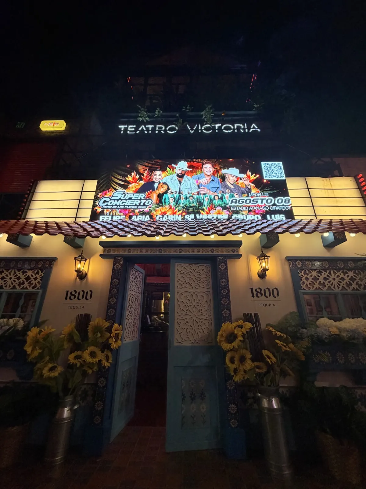 The entrance of Teatro Victoria at night, with its lit sign and a screen above blue doors and pots of sunflowers