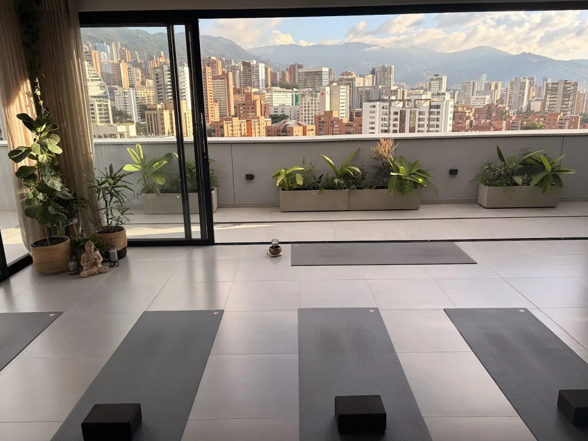 The YOGALAB studio with yoga mats and blocks laid out on a pale tiled floor, glass doors open onto a planted terrace and a view of El Poblado's towers and the mountains